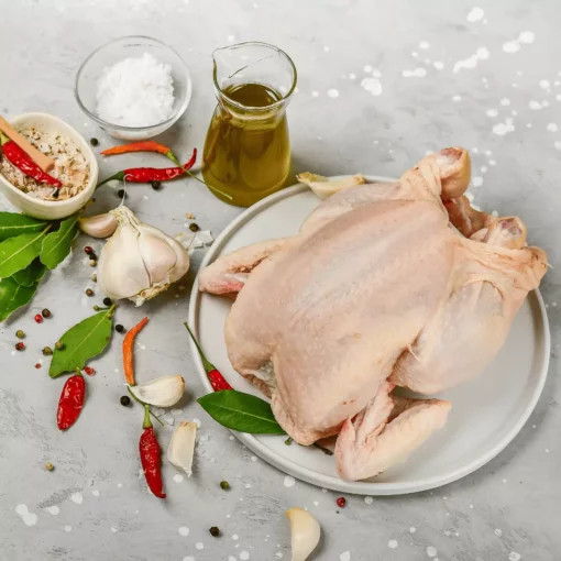 Une table blanche/grise sur laquelle il y a une assiette blanche qui contient un poulet entier blanc, de l'huile d'olive, des feuilles de laurier, de l'ail, du piment rouge et du gros sel